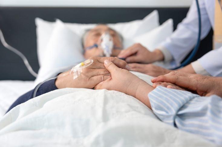 Woman holds the hand of an elderly nursing home resident being examined by a healthcare professional.  